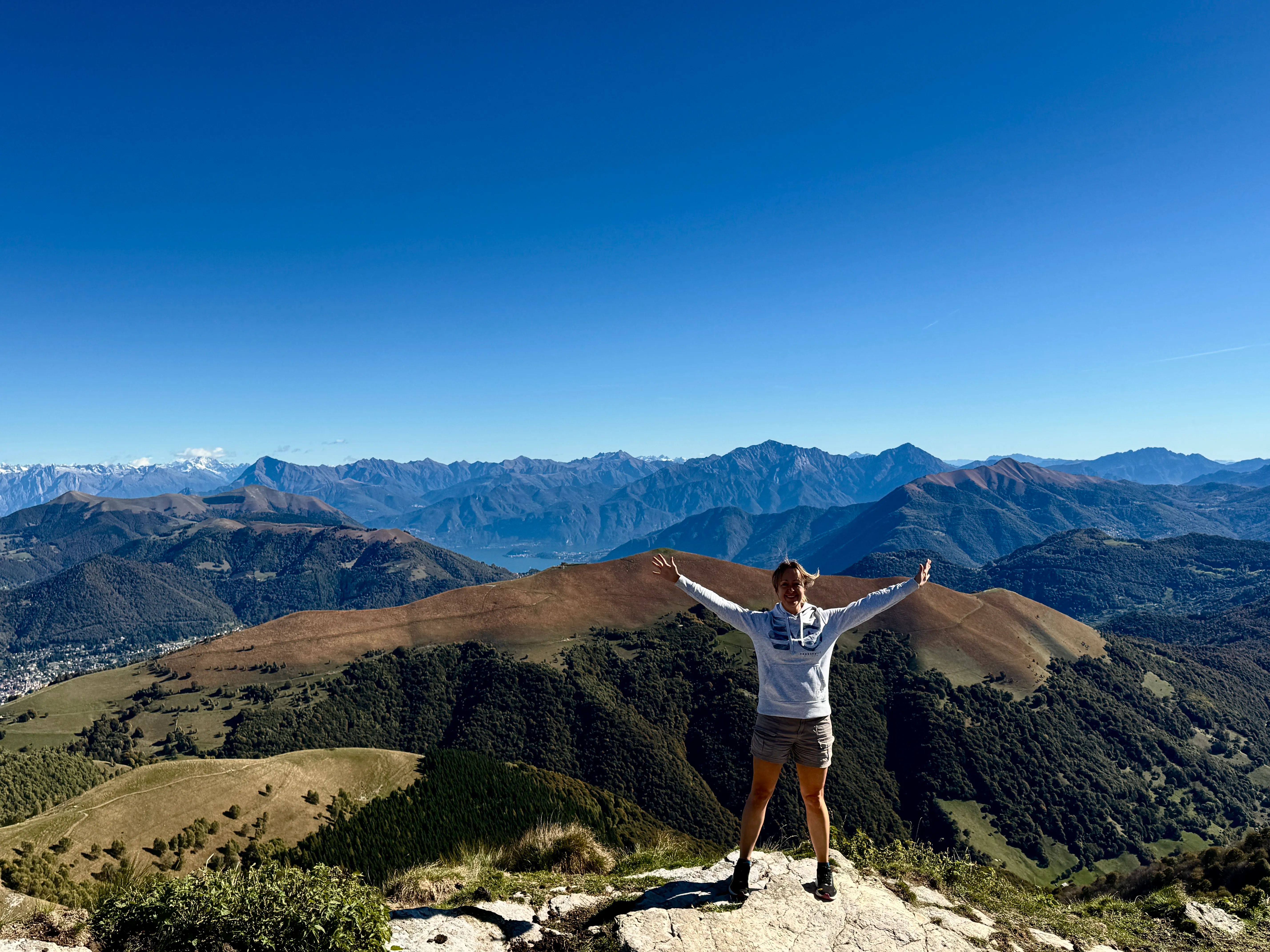 Becky with arms outstretched at the summit of Monte Generoso, with Lake Lugano visible far below