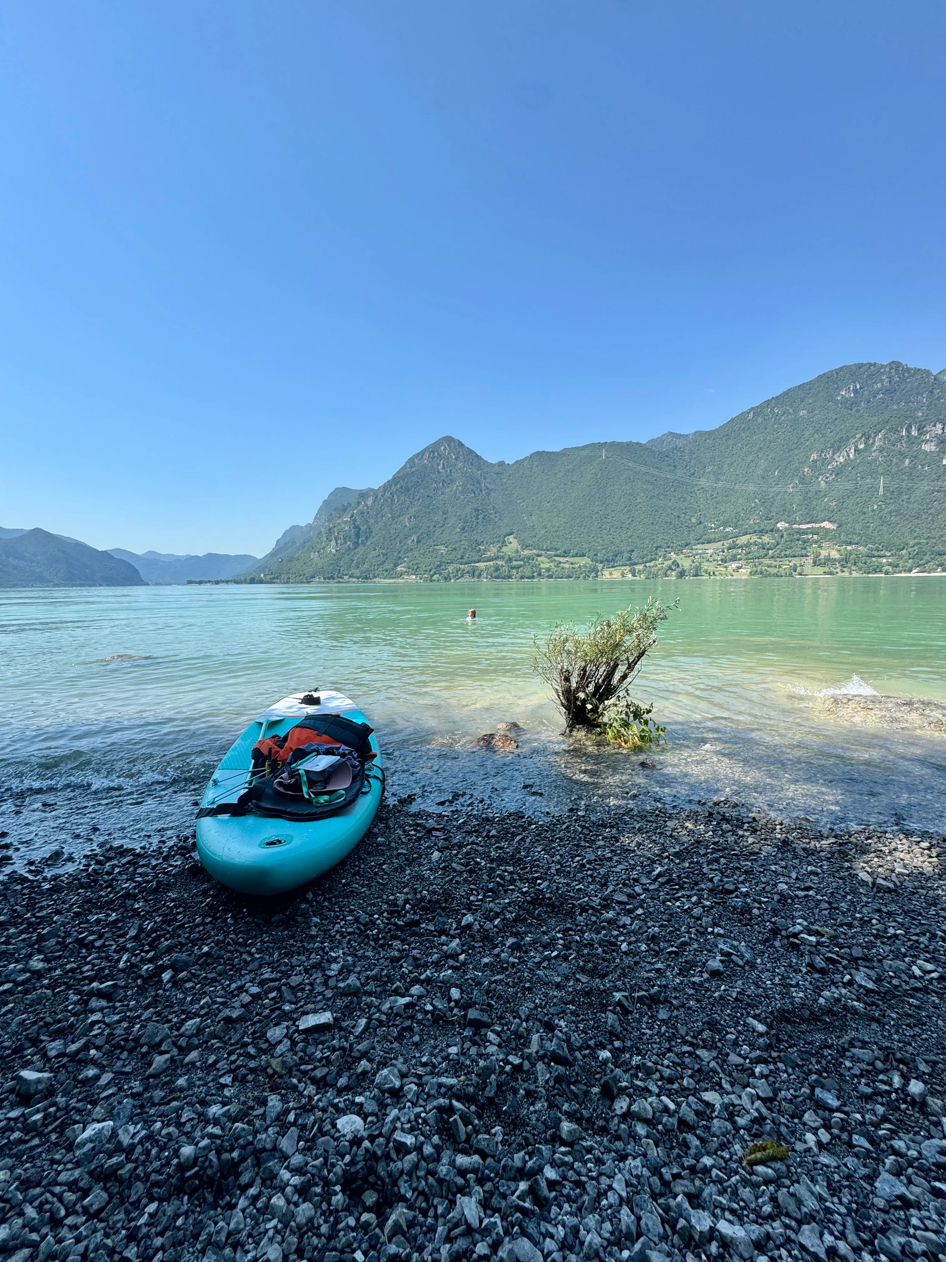 A paddleboard resting on a quiet pebble beach on Lago d’Idro with someone swimming in the turquoise water