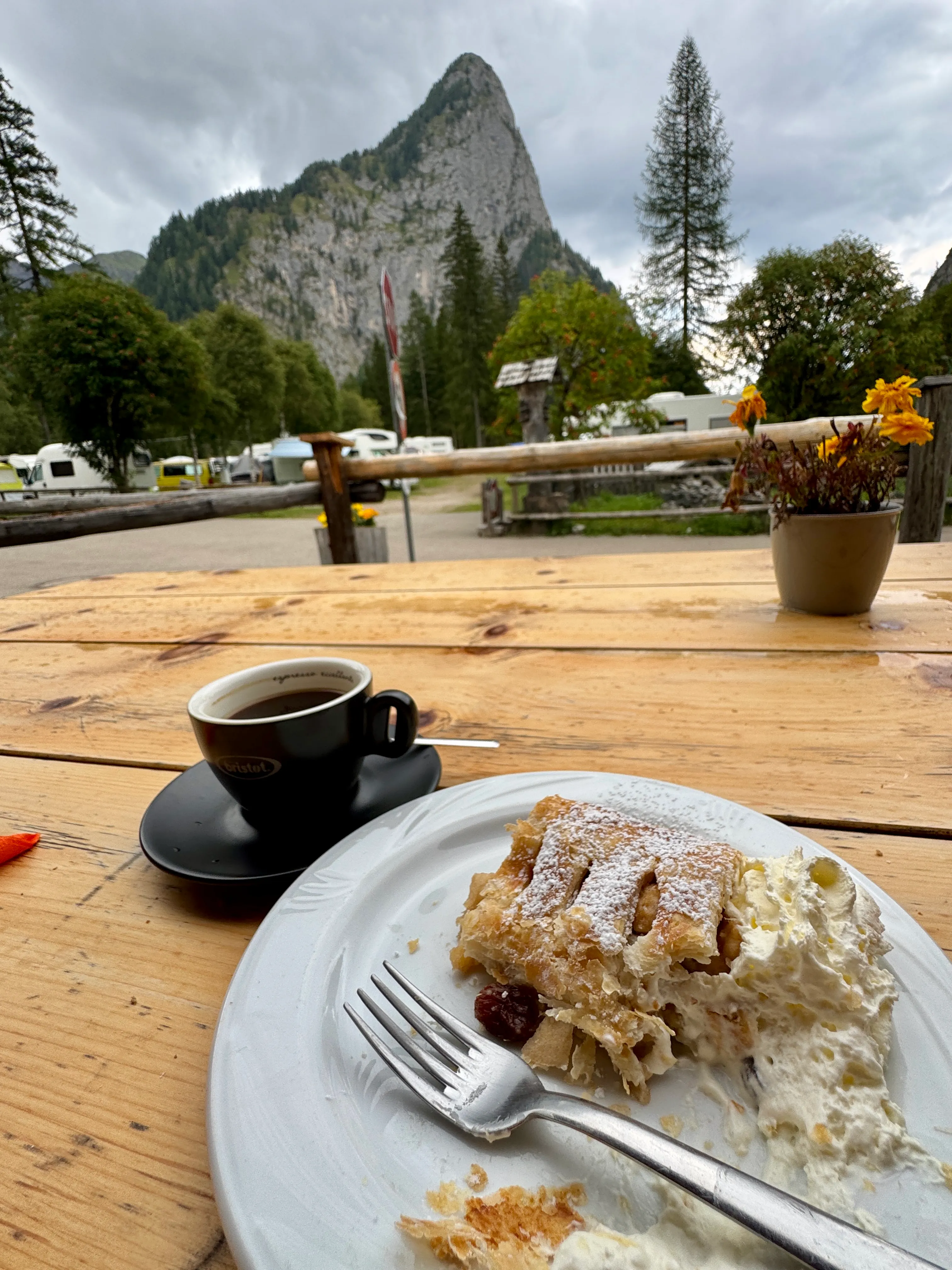 A slice of homemade apple strudel with cream and an espresso on a wooden table, mountains behind